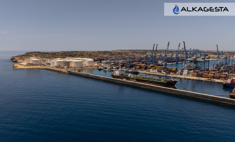 Aerial view of a coastal industrial port with cargo cranes, storage tanks, and docked vessels, featuring the Alkagesta logo at the top.