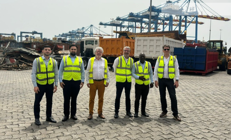 Group of individuals wearing safety vests standing at an industrial port area with cargo containers and cranes during Alkagesta’s West Africa visit.