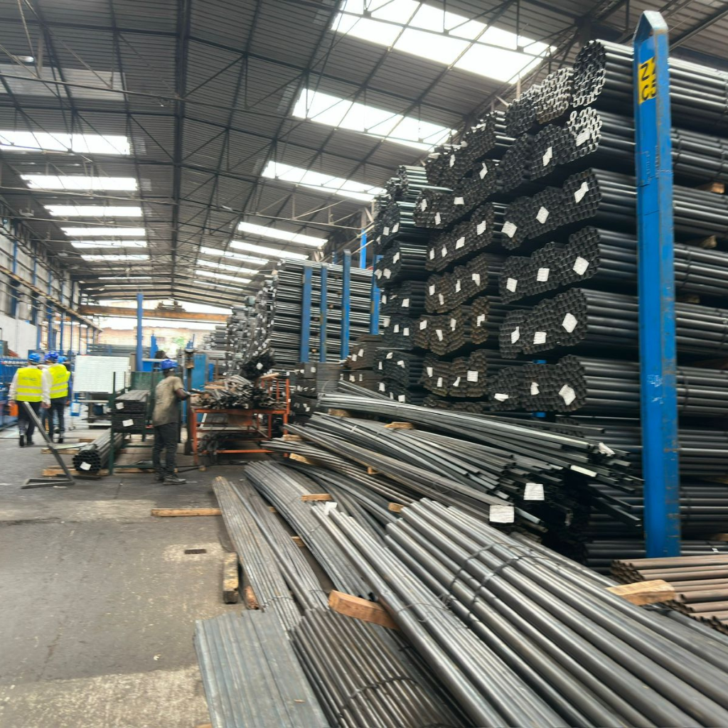 Industrial warehouse with long stacked metal rods arranged in tall racks and individuals wearing safety vests inspecting materials during Alkagesta’s West Africa site visit.