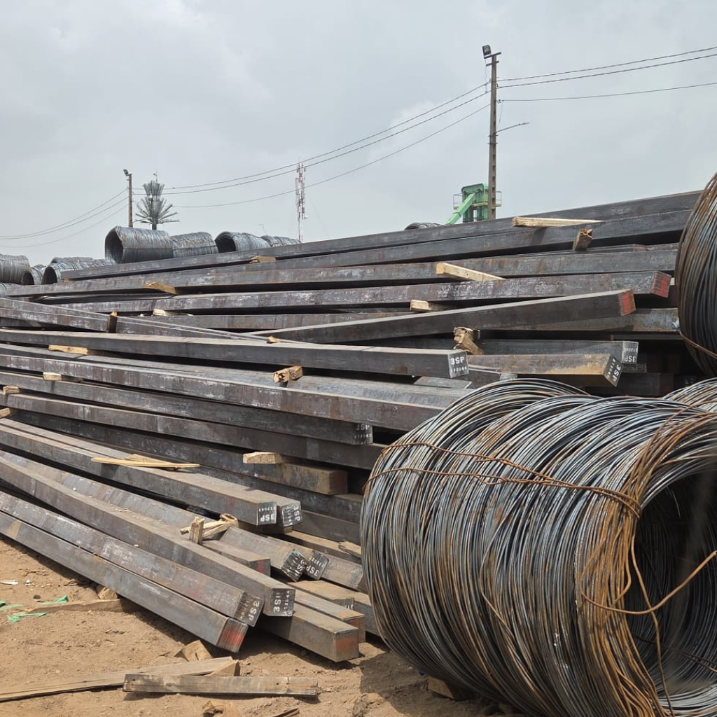 Stacks of long metal beams and a large industrial wire coil stored outdoors at a construction materials yard during Alkagesta’s West Africa site visit.