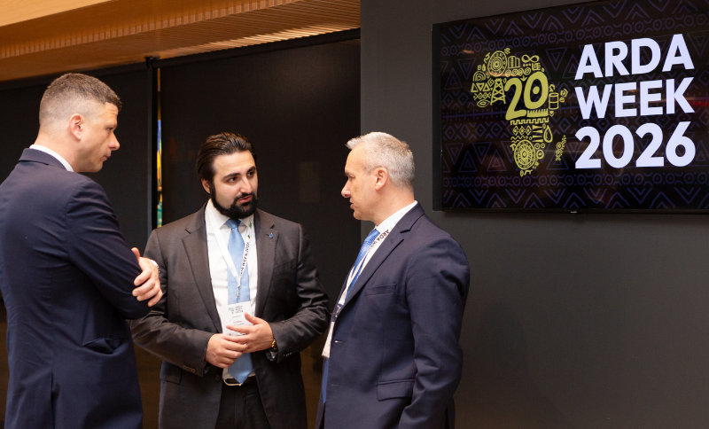 Three conference participants standing and talking in front of a display reading “ARDA Week 2026” during an indoor industry event.