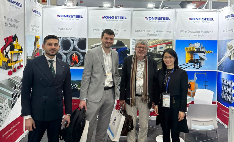 Representatives standing at the WONGSTEEL exhibition stand at Wire & Tube Düsseldorf, with product banners for wire rod machinery, spheroidizing annealing furnaces, and wire drawing machines visible in the background.