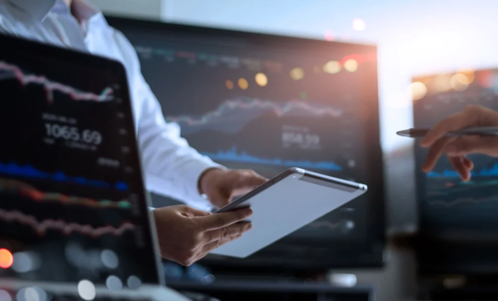 Two people reviewing financial and performance charts displayed on large screens, with one person holding a tablet and another pointing at data during an analytical discussion.
