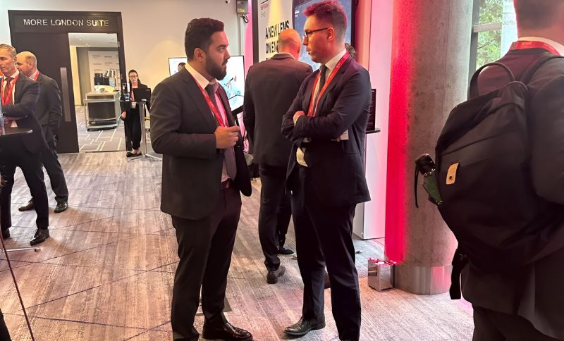 Two men in suits with red lanyards talk in a modern conference area, surrounded by other attendees. A large concrete pillar and a sign reading MORE LONDON SUITE are visible in the background.