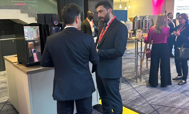Several people in business attire are gathered around a coffee station at a professional event, engaging in conversation. Two men in the foreground are talking near a mini fridge and coffee cups.