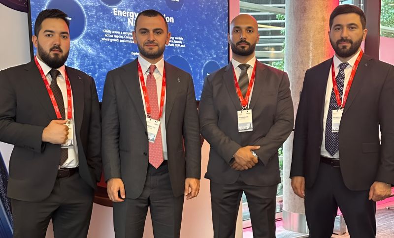 Four men in suits with red lanyards and name badges stand indoors in front of a screen displaying text about energy. The background includes a window and pillars.