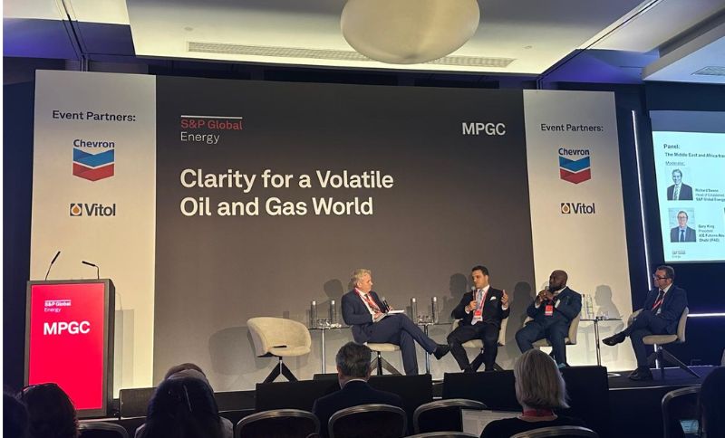 Four men sit on a stage in a panel discussion at an energy conference titled Clarity for a Volatile Oil and Gas World, with event partner logos for Chevron and Vitol displayed on banners. Audience members are visible in front.