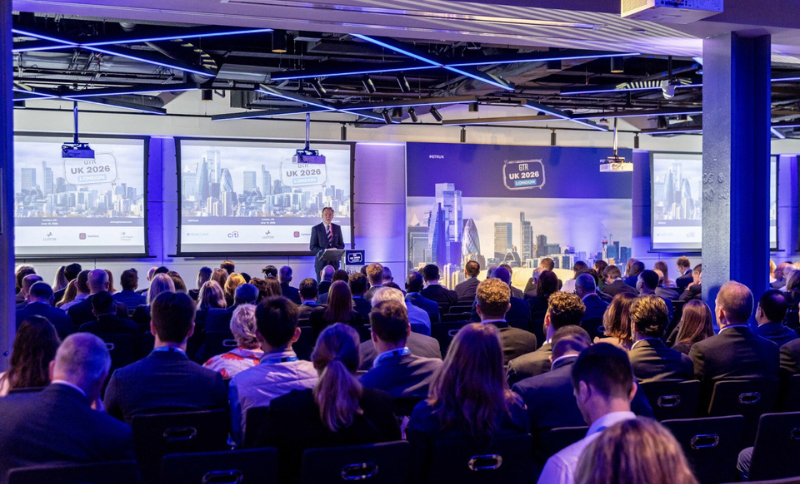 A large audience sits in rows facing a stage where a speaker presents at a conference. Two large screens display UK 2026 with images of a city skyline in the background. The room is modern and well-lit.