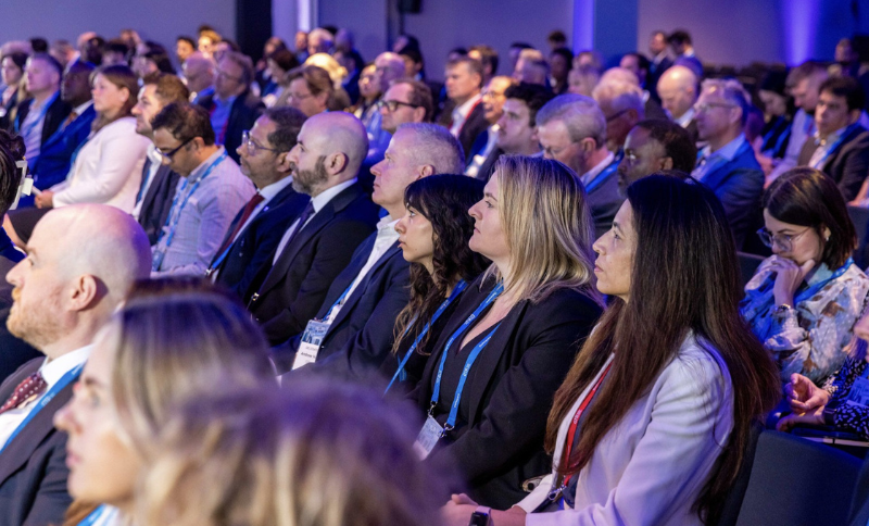 A diverse group of people in business attire sit attentively in rows at a conference or seminar, with blue lighting highlighting the focused audience members.