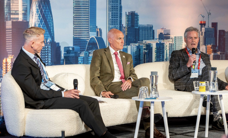 Three men in suits sit on a white sofa, participating in a panel discussion. One holds a microphone while speaking. Modern city skyscrapers are visible in the background.