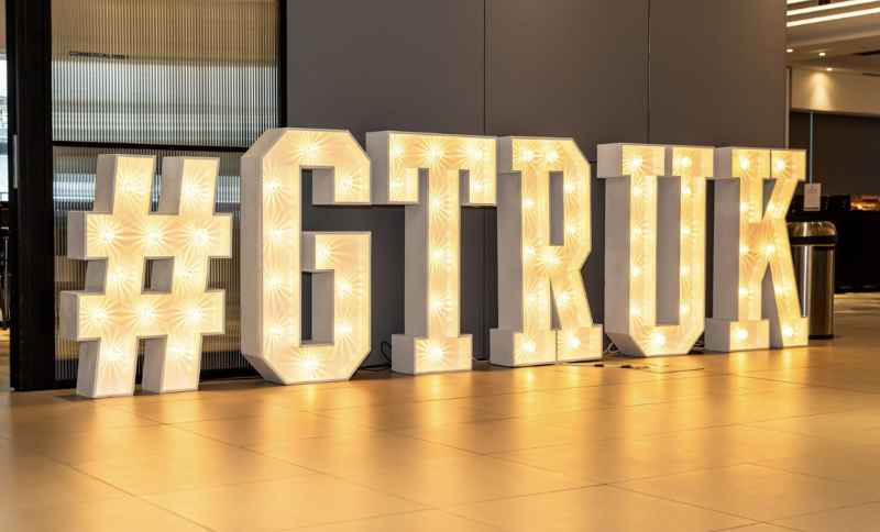 Large, illuminated marquee letters and a hash symbol spell out “#GTRUK” in a modern indoor setting with tiled floors and a neutral-coloured wall in the background.
