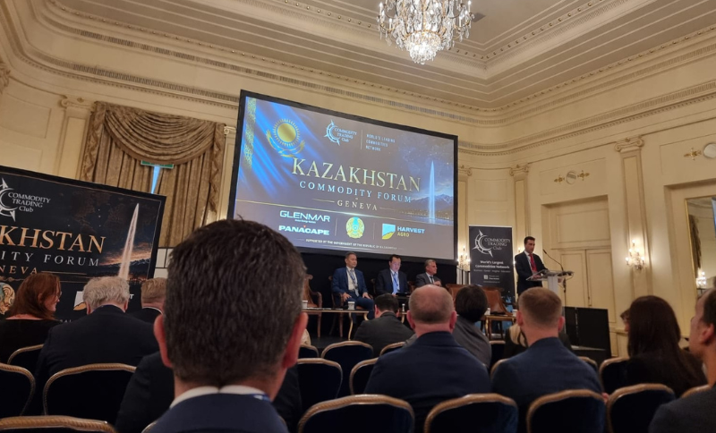 A conference room with attendees seated, facing a stage where five men are participating in the Kazakhstan Commodity Forum in Geneva. One man is speaking at a lectern, and a large screen displays event and sponsor logos.