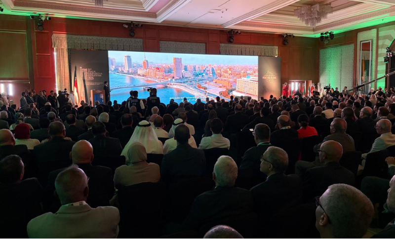 A large audience in formal attire sits facing a stage with a wide screen displaying a cityscape in a well-lit conference hall. Some attendees wear traditional Middle Eastern clothing.