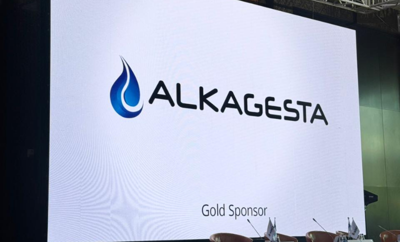 A large screen displays the ALKAGESTA logo with a blue drop symbol and the words Gold Sponsor below it; chairs and microphones are set up in front, suggesting a conference or event setting.