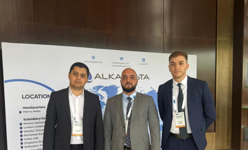 Three men in business attire stand in front of a banner displaying company information and a world map. All are wearing conference name badges and looking at the camera, with a wood-panelled wall in the background.