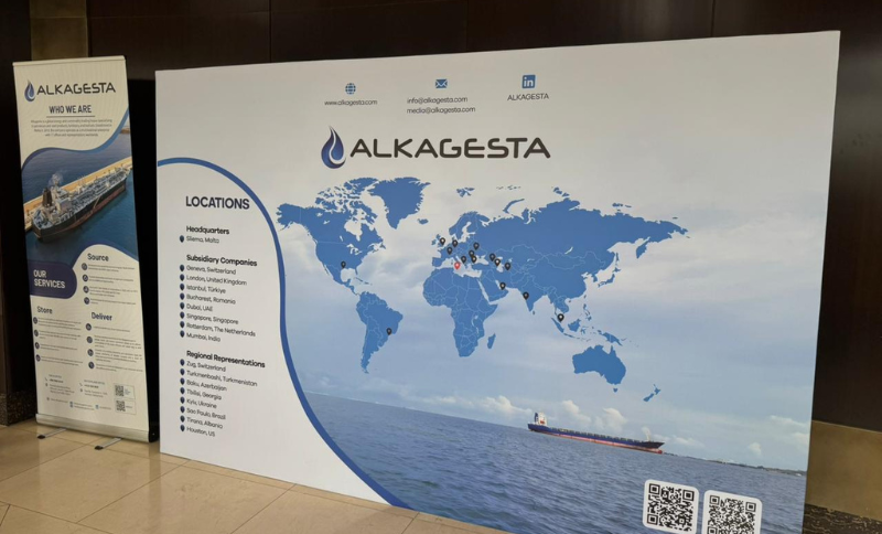 Large display board showing the ALKAGESTA logo, a world map with marked locations, company services, contact information, and a ship at sea along the bottom. Subsidiary and regional offices are listed on the left.