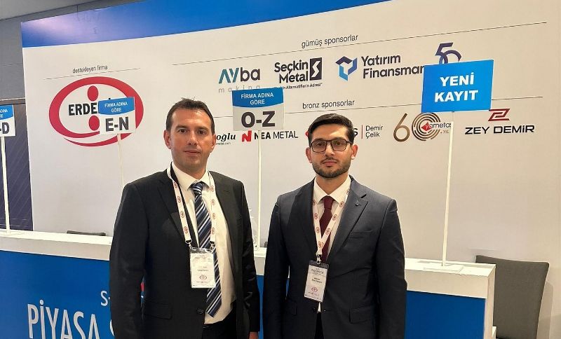 Two men in suits stand in front of a registration desk at an event, with sponsorship banners displayed on the wall behind them. One holds a sign reading Yeni Kayıt, which means New Registration in Turkish.