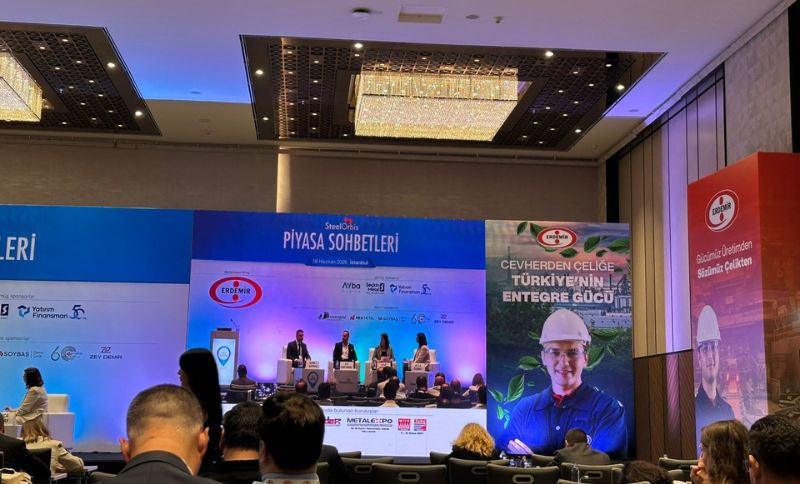 A conference panel with five speakers sits on a stage before an audience in a modern hall. The backdrop displays Turkish text, logos, and a large image of a man in safety gear promoting Turkish steel.