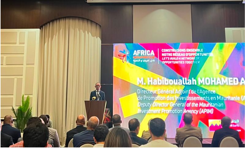 A man stands at a lectern speaking to an audience in a conference room. Behind him is a large screen displaying event branding and speaker information for M. Habiboullah Mohamed Abed, Deputy Director General of APIM.