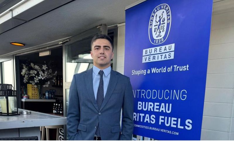 A man in a suit stands indoors next to a blue Bureau Veritas banner that reads Shaping a World of Trust and Introducing Bureau Veritas Fuels. There are flowers and shelves in the background.