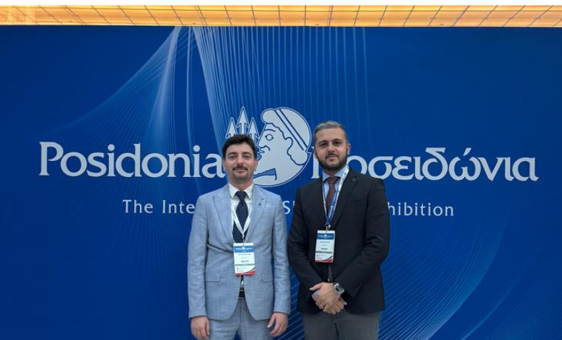 Two men in business attire stand in front of a blue backdrop with Posidonia and Greek text, at the International Shipping Exhibition. Both wear name badges and look at the camera.