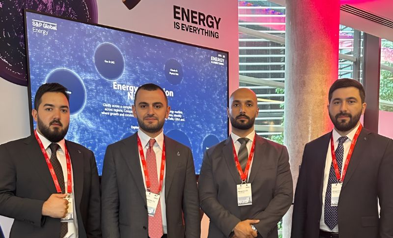 Four men in business suits with red lanyards stand in front of a digital display about energy at a professional event. A sign behind them reads ENERGY IS EVERYTHING.