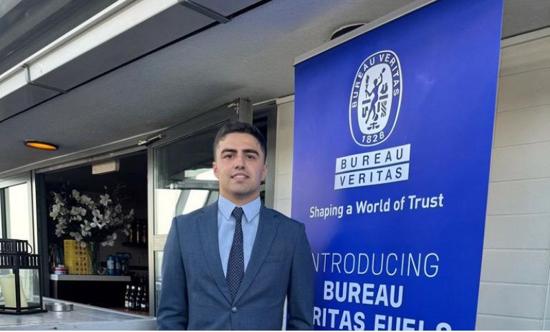 A man in a suit stands next to a blue Bureau Veritas banner that reads Shaping a World of Trust and Introducing Bureau Veritas Fuel. The setting appears to be an indoor-outdoor event space.
