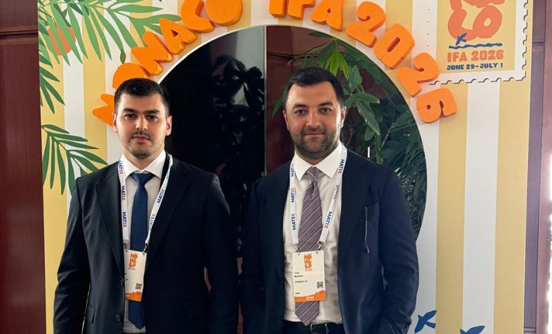 Two men in suits with conference badges stand in front of a decorative arch reading Monaco IFA 2026, with tropical-themed decor and event details in the background.