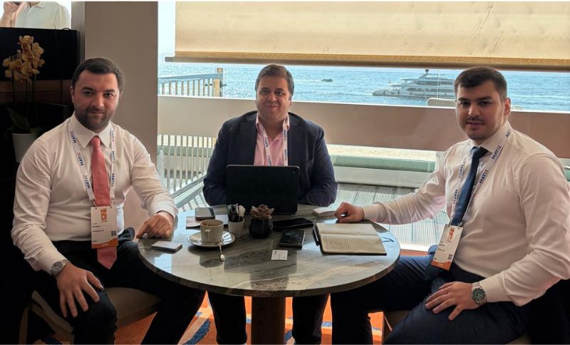 Three men in business attire sit around a small round table with laptops, papers, and coffee cups, in front of a large window overlooking a marina with boats and water in the background.