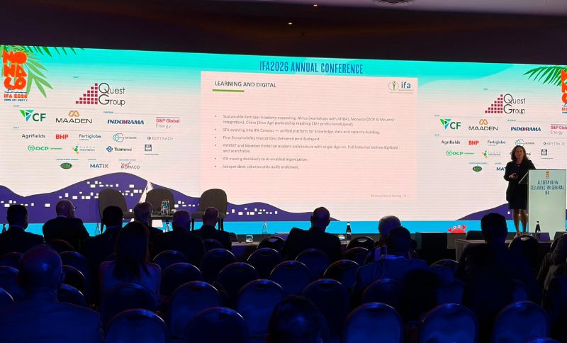 A conference room with people seated facing a large screen displaying a presentation. The slide is titled LEARNING AND DIGITAL, and logos of various sponsors are visible at the bottom of the screen. A speaker stands at a lectern on the right.