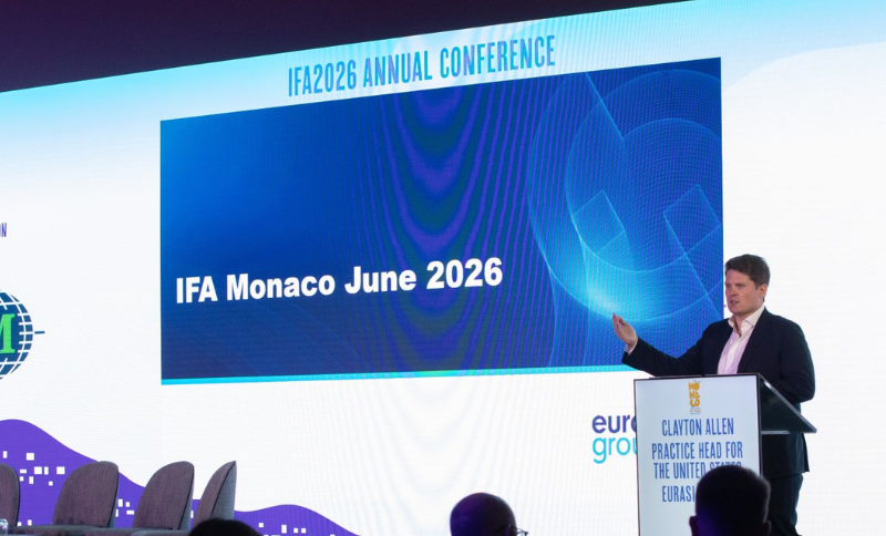 A man in a suit speaks on stage beside a large screen displaying “IFA Monaco June 2026” at the IFA2026 Annual Conference. There are empty chairs and a few audience members visible in the foreground.