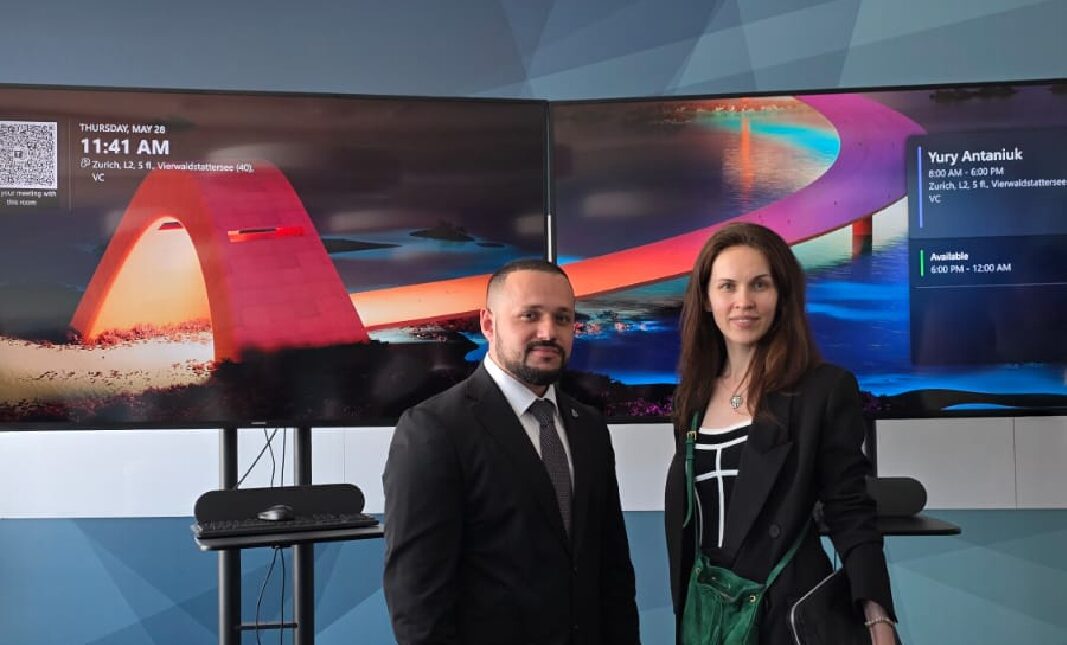 Two people in business attire stand in front of large monitors displaying a colourful bridge image and digital meeting details.