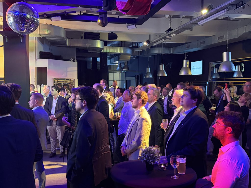 A large group of people in business attire stand attentively at an indoor event with colourful lighting, a disco ball above, and drinks on nearby tables.
