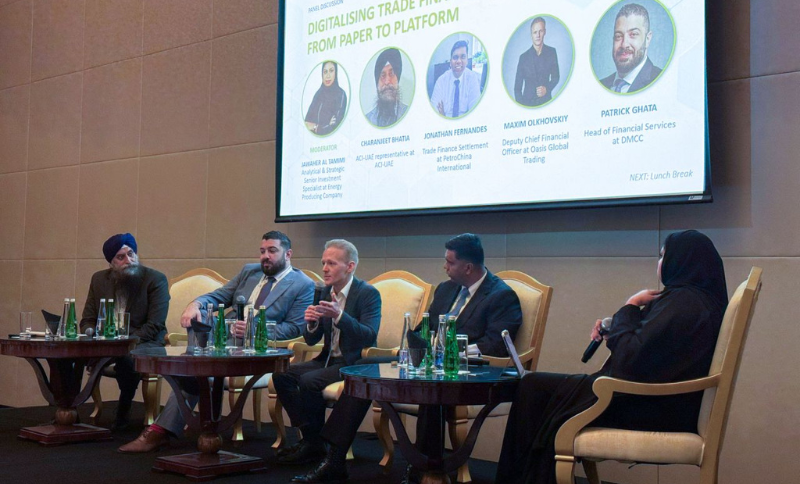 Five panellists sit on a stage, engaged in a discussion. A large screen behind them displays their names and photos, and water bottles are placed on the table in front. The setting appears to be a formal conference or seminar.