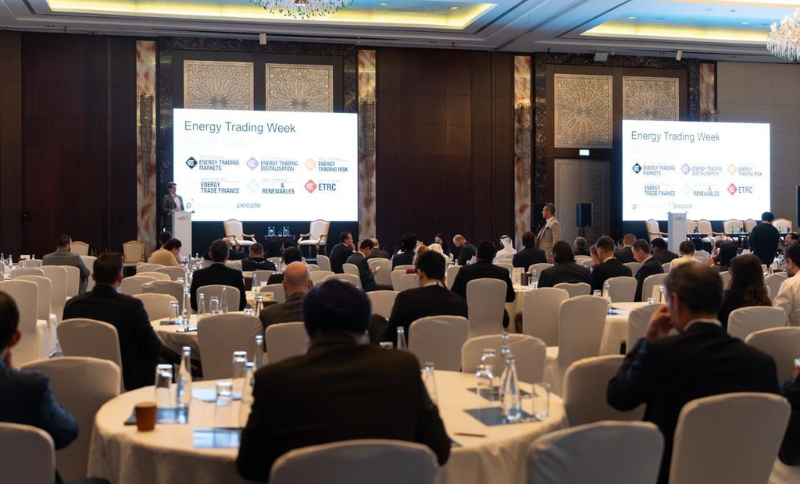 Business conference with attendees seated at round tables, facing two large screens displaying Energy Trading Week and sponsor logos. The room has elegant décor and soft lighting.