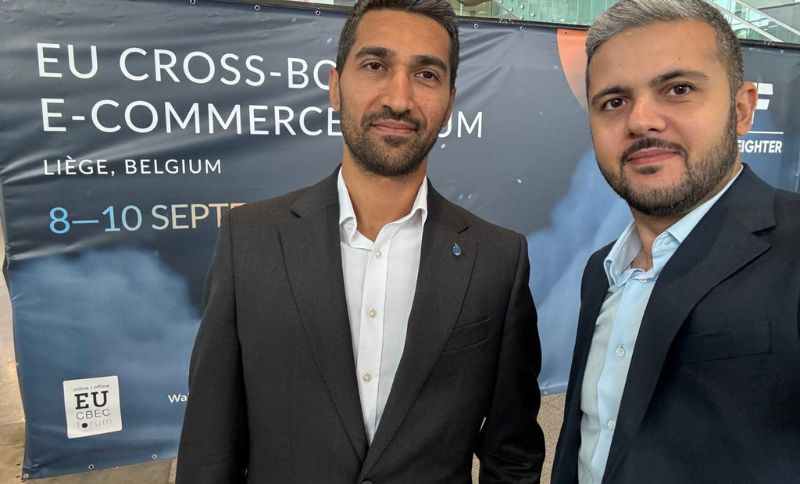 Two men in suits take a selfie in front of a banner for the EU Cross-Border E-Commerce Forum in Liège, Belgium, scheduled for 8–10 September.