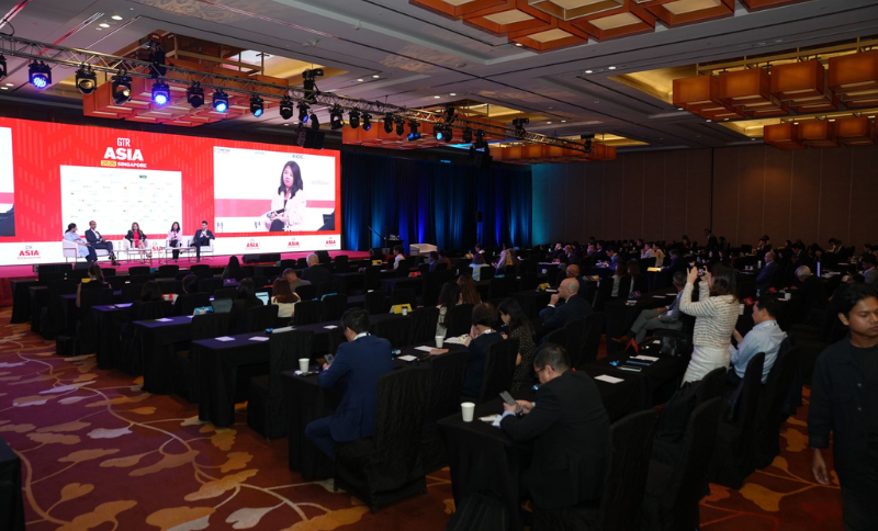 Attendees sit at tables in a large conference room, watching panellists onstage with a large FT Asia screen behind them. The room is dimly lit with spotlights on the stage.