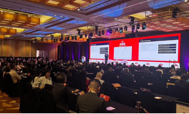 A speaker presents slides to a large audience in a conference hall with red Asia branding on screens, rows of seated attendees, and elaborate ceiling lights.