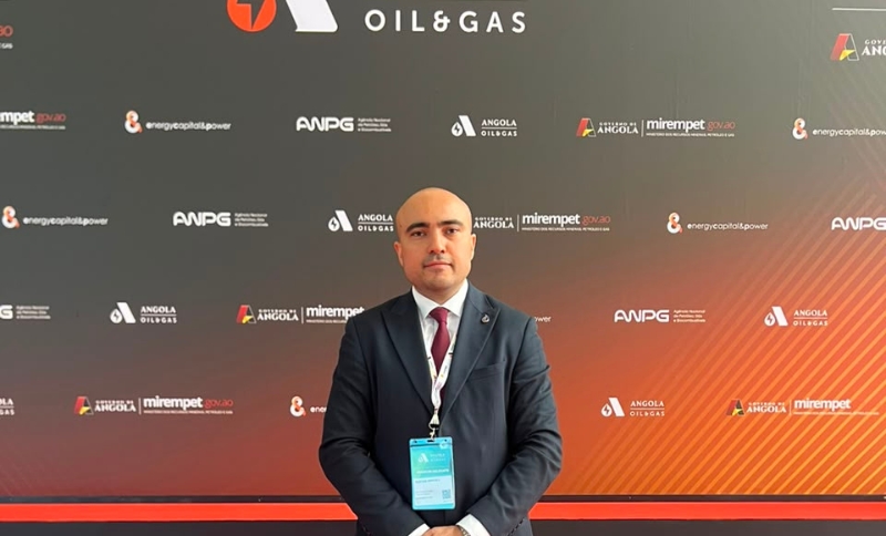 A man in a suit stands in front of a backdrop with oil and gas logos at a conference. He wears a lanyard and badge, facing the camera with a neutral expression. The background features event sponsor names and logos.