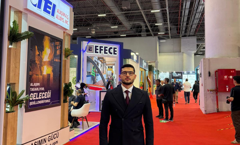 A man in a suit stands in a brightly lit exhibition hall with red carpet, surrounded by stands and people walking or sitting.