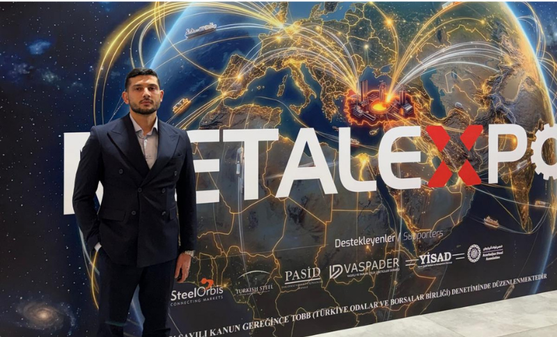 A man in a dark suit stands in front of a large METAL EXPO sign with a world map and city lights in the background. Various sponsor logos appear along the bottom of the display.