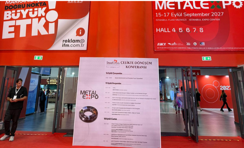 A large stand at a trade exhibition displays conference information on a central board. Red banners above show METAL EXPO and the event dates. Two people stand near the entrance, and several exhibition halls are visible inside.