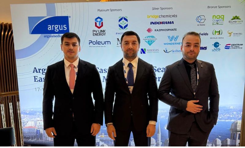 Three men in suits stand in front of a conference backdrop displaying various sponsor logos, including Argus, PV Link Energy, and Kazphosphate. The event appears to be business-related.
