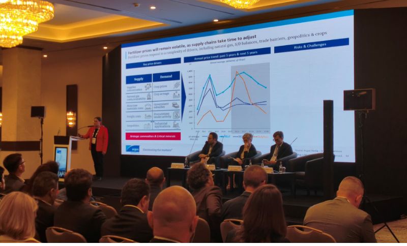 A speaker presents to an audience in a conference room, with a large screen displaying supply chain graphs and data. Three panellists are seated on stage, and attendees are watching attentively.