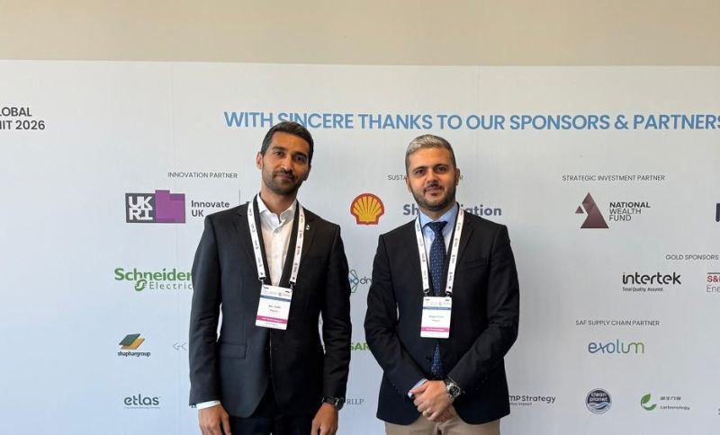 Two men in business attire stand in front of a sponsor backdrop at a conference or summit, both wearing lanyards and looking at the camera, with various sponsor logos visible behind them.