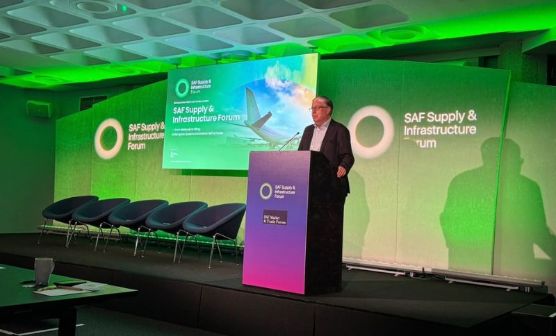 A speaker stands at a lectern on stage at the SAF Supply & Infrastructure Forum, with green-lit walls, empty chairs, and a large presentation screen showing an aeroplane image in the background.