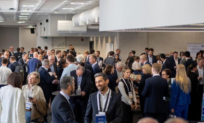 A large group of people in business attire mingle and converse at a busy indoor networking event or conference, with tables scattered around the room.
