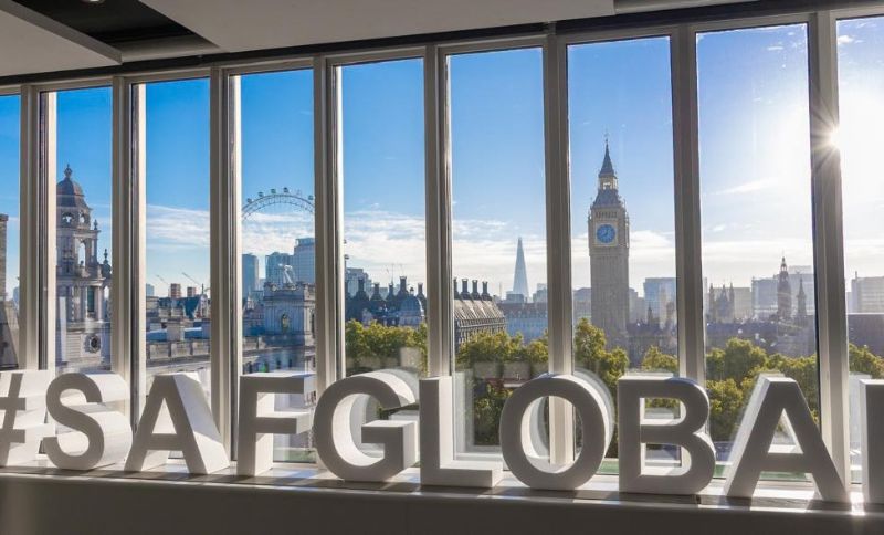Large white letters spell #SAFGLOBAL indoors, with tall windows showing a clear view of London landmarks, including the London Eye and Big Ben, under a bright blue sky.