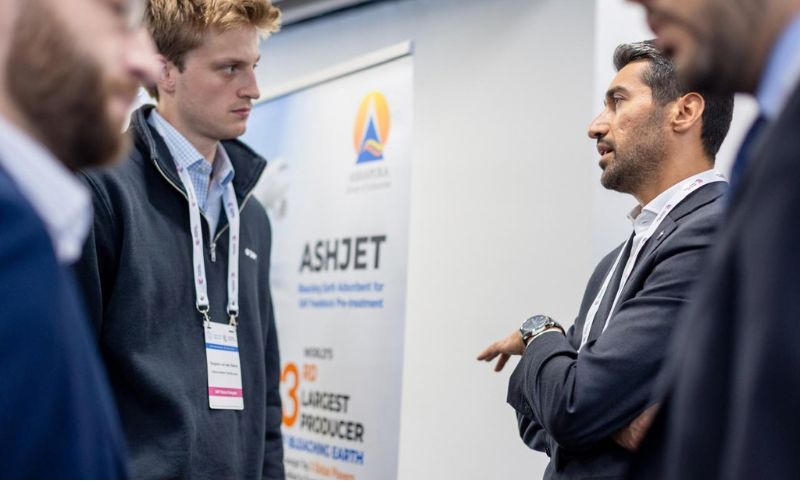 Three men stand in conversation at a professional event. One man gestures with his hands. A sign behind them displays ASHJET and mentions world’s 3rd largest producer. The atmosphere is businesslike.