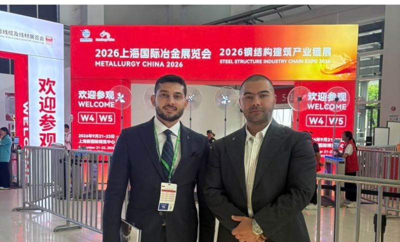 Two men in business attire stand inside a convention centre with red signs behind them for Metallurgy China 2026 and Steel Structure Industry Chain Expo 2026. They are posing for a photo near event entrance barriers.